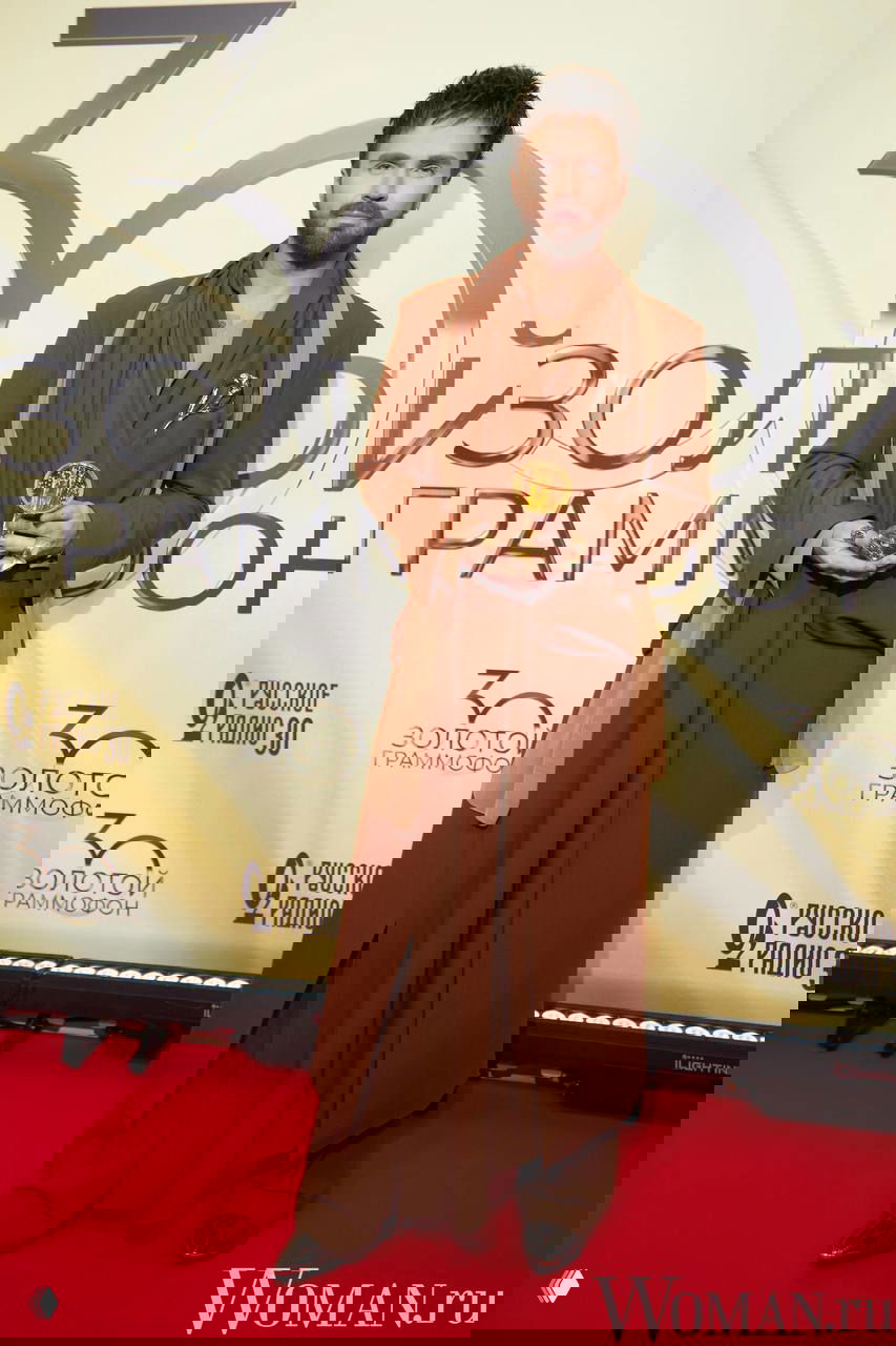 Александр Панайотов в коричневом многослойном образе и шарфе держит награду на красной дорожке, на фоне крупные элементы фирменного оформления события.