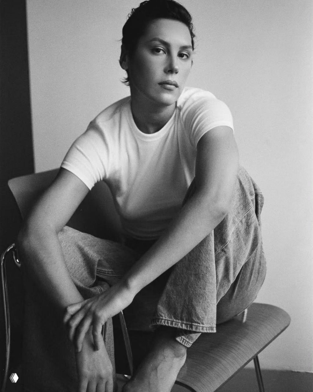 Чёрно‑белый портрет женщины в белой футболке и джинсах, сидящая на стуле в студии с мягким естественным светом.