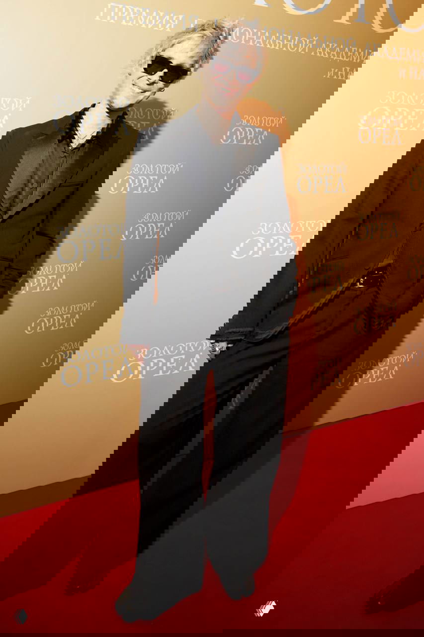 Мужчина в стильном чёрном наряде и солнечных очках улыбается на красной дорожке премии «Золотой орел»; неформальный вечерний лук.