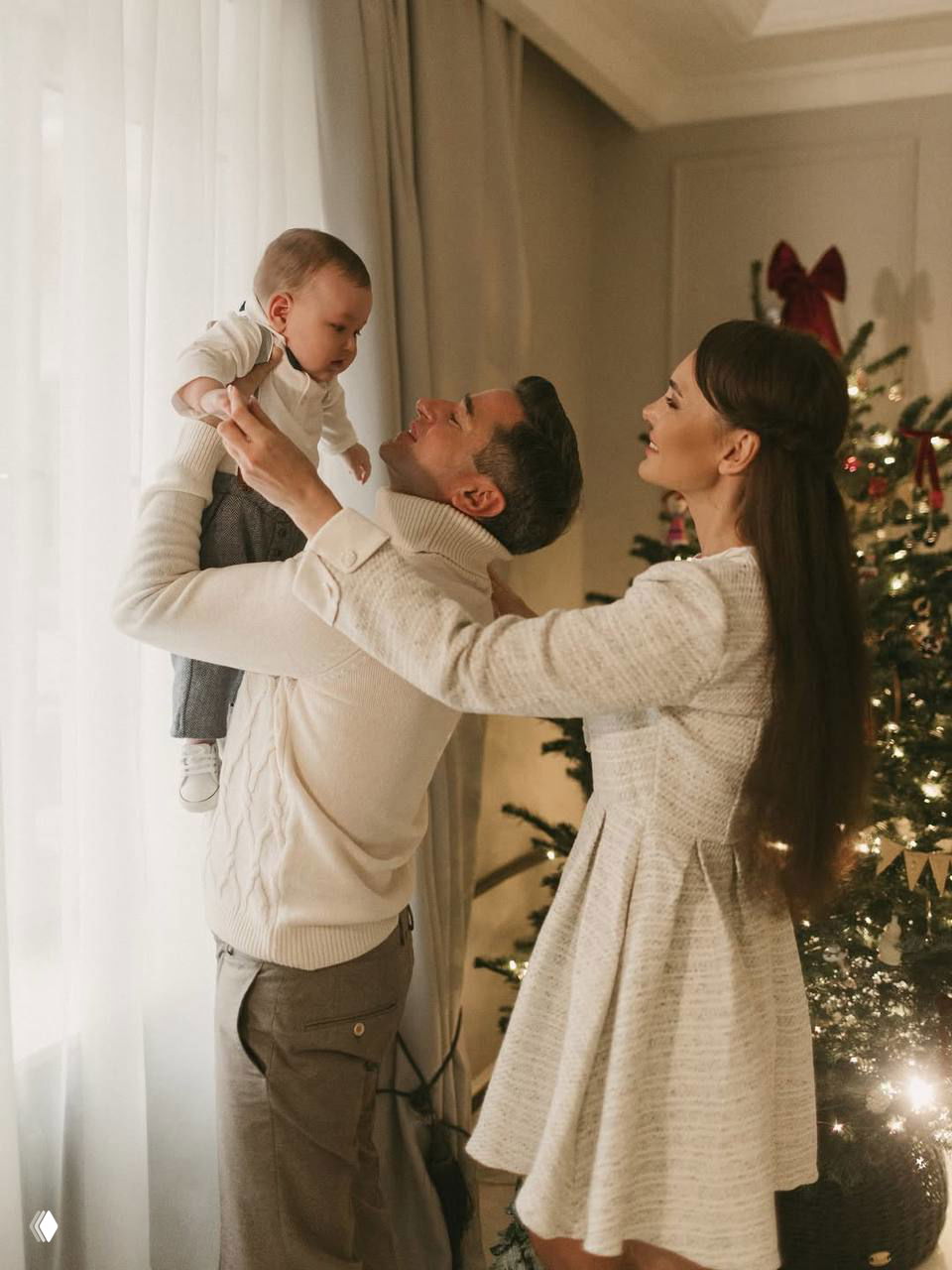 Мужчина поднимает малыша в руках у окна на фоне наряженной ёлки — семейный портрет с мягким светом и тёплой атмосферой.