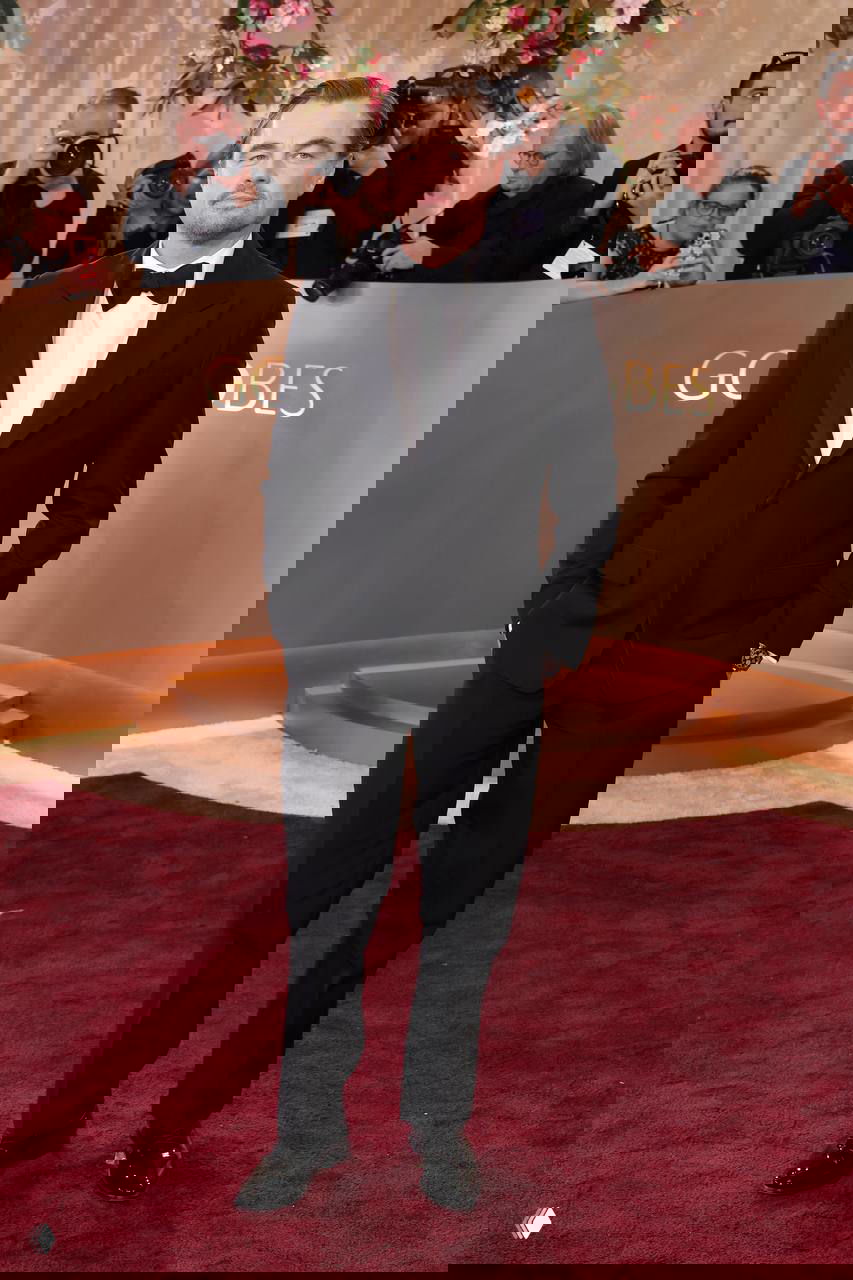 Мужчина в строгом черном костюме и бабочке позирует у декораций церемонии «Золотой глобус», вокруг видны фотографы и пресса.