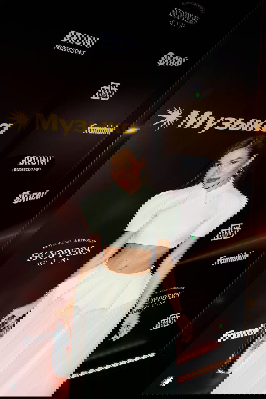 Женщина в белом укороченном топе и многослойной юбке позирует на пресс‑волле премии с логотипом «Яндекс Музыка» под вспышки камер.