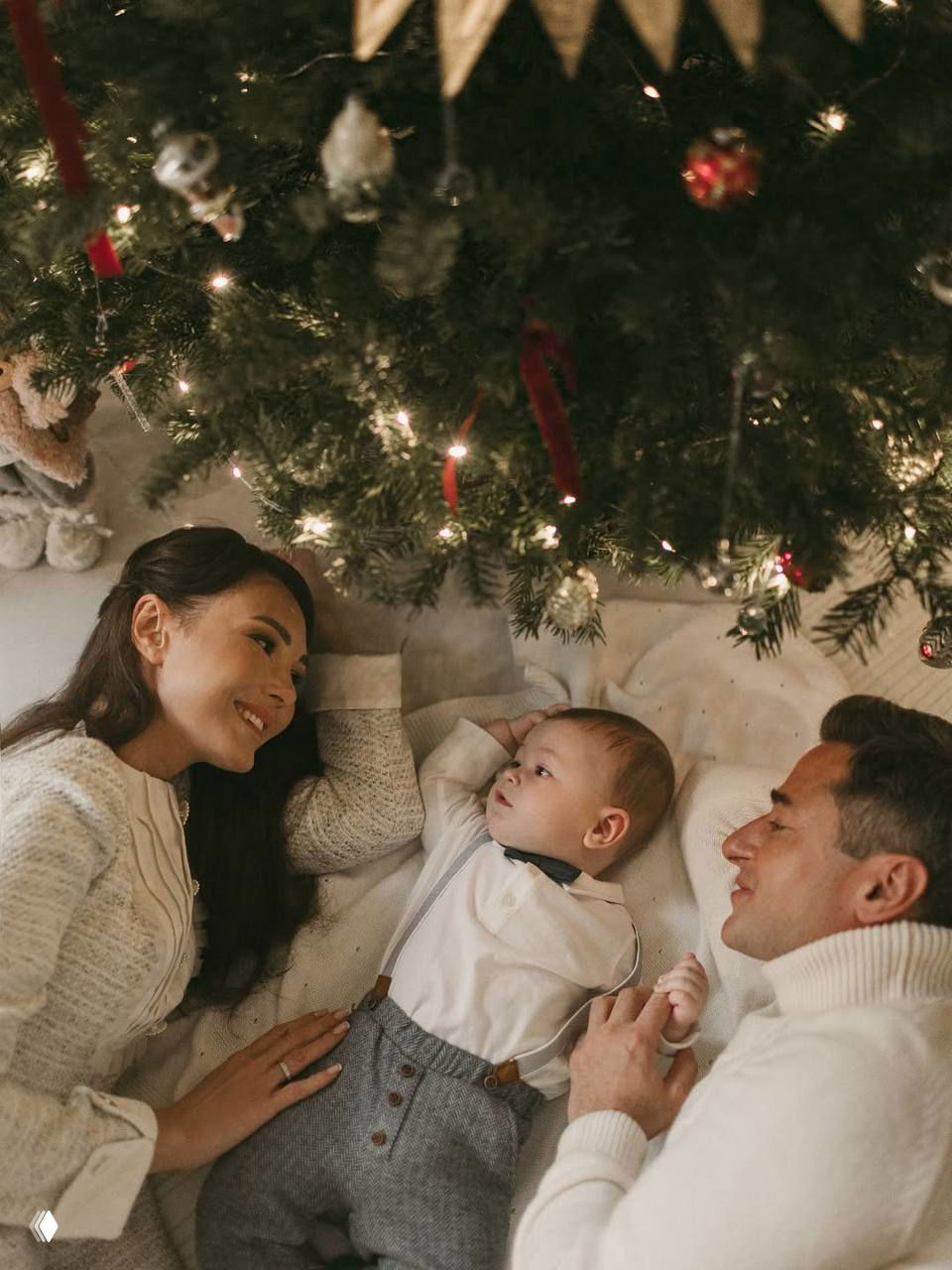 Женщина, мужчина и ребёнок лежат у ёлки и смотрят друг на друга; кадр передаёт спокойствие семейного праздника и нежность взаимоотношений.