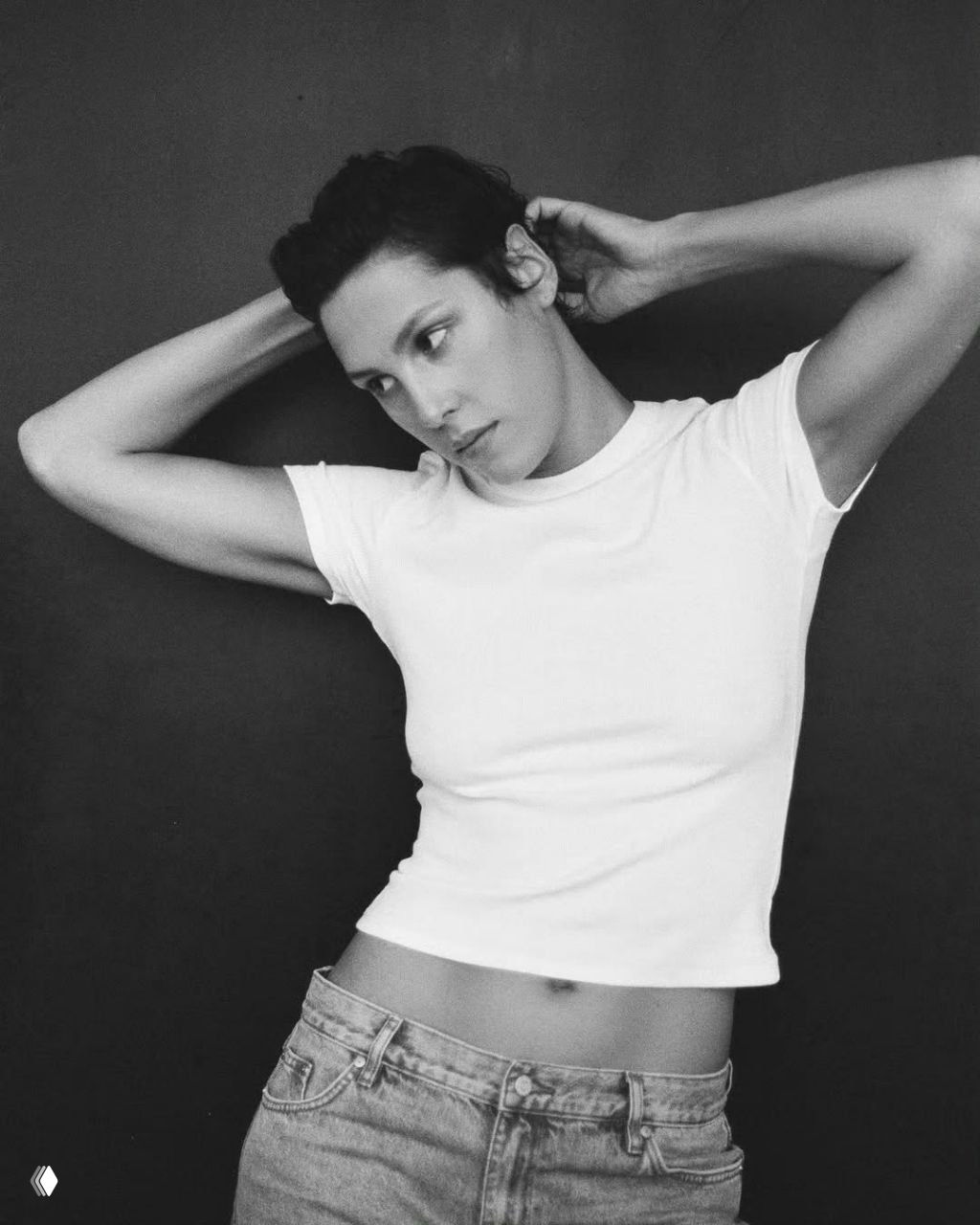 Чёрно‑белый портрет: женщина с короткими волосами поправляет причёску, белая футболка и джинсы на тёмном фоне.