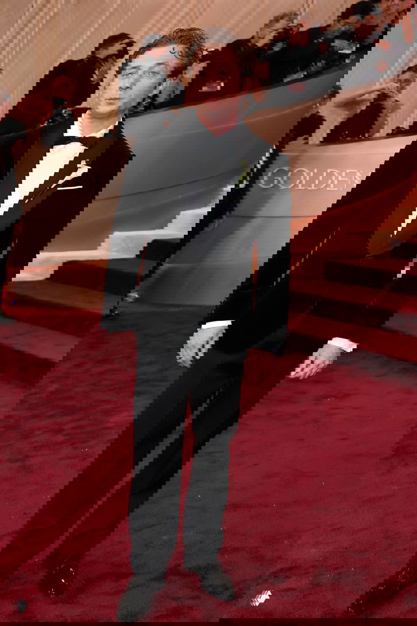 Мужчина в бархатном смокинге с бутоньеркой стоит на красной дорожке «Золотого глобуса», в кадре — профессиональные фотокамеры.