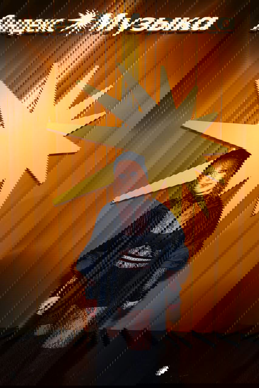 Рыжеволосая женщина в прозрачном кружевном наряде и оверсайз‑куртке позирует у золотой инсталляции с символикой премии «Яндекс Музыка».