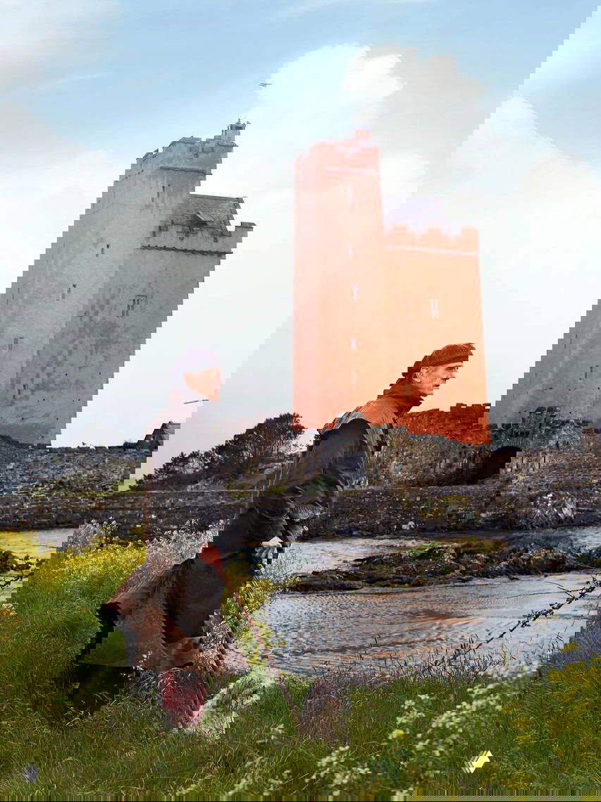 Внешний вид красочной башни замка Килко на фоне неба; на переднем плане Джереми Айронс стоит с прогулочной палкой и собакой у береговой линии.