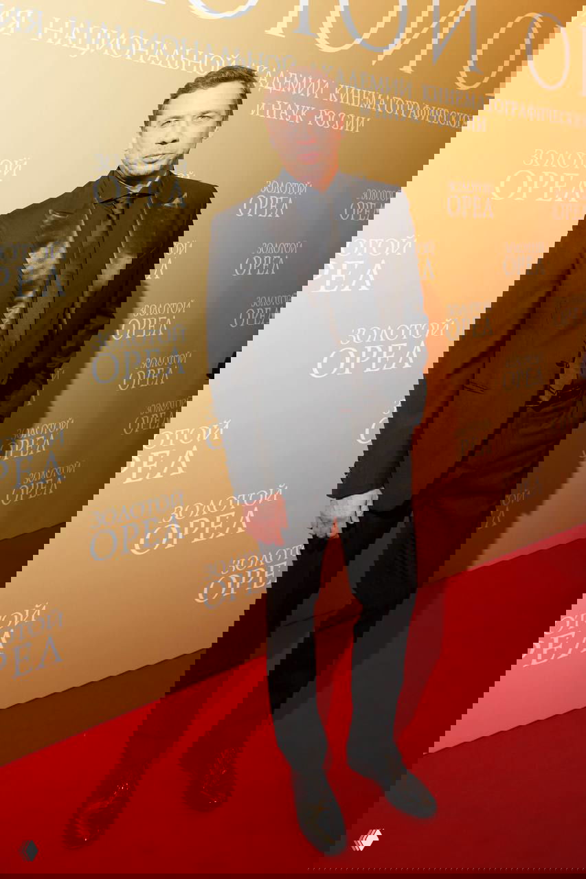 Мужчина в чёрном костюме и тёмном галстуке стоит на красной дорожке у золотого баннера премии «Золотой орел», официальный портретный кадр.