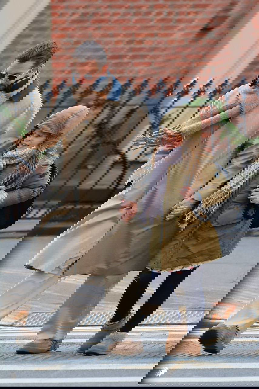 Мужчина в бежевом длинном плаще, шарфе и наушниках идёт по тротуару, держа за руку ребёнка в тёплой зелёной куртке и шарфе.