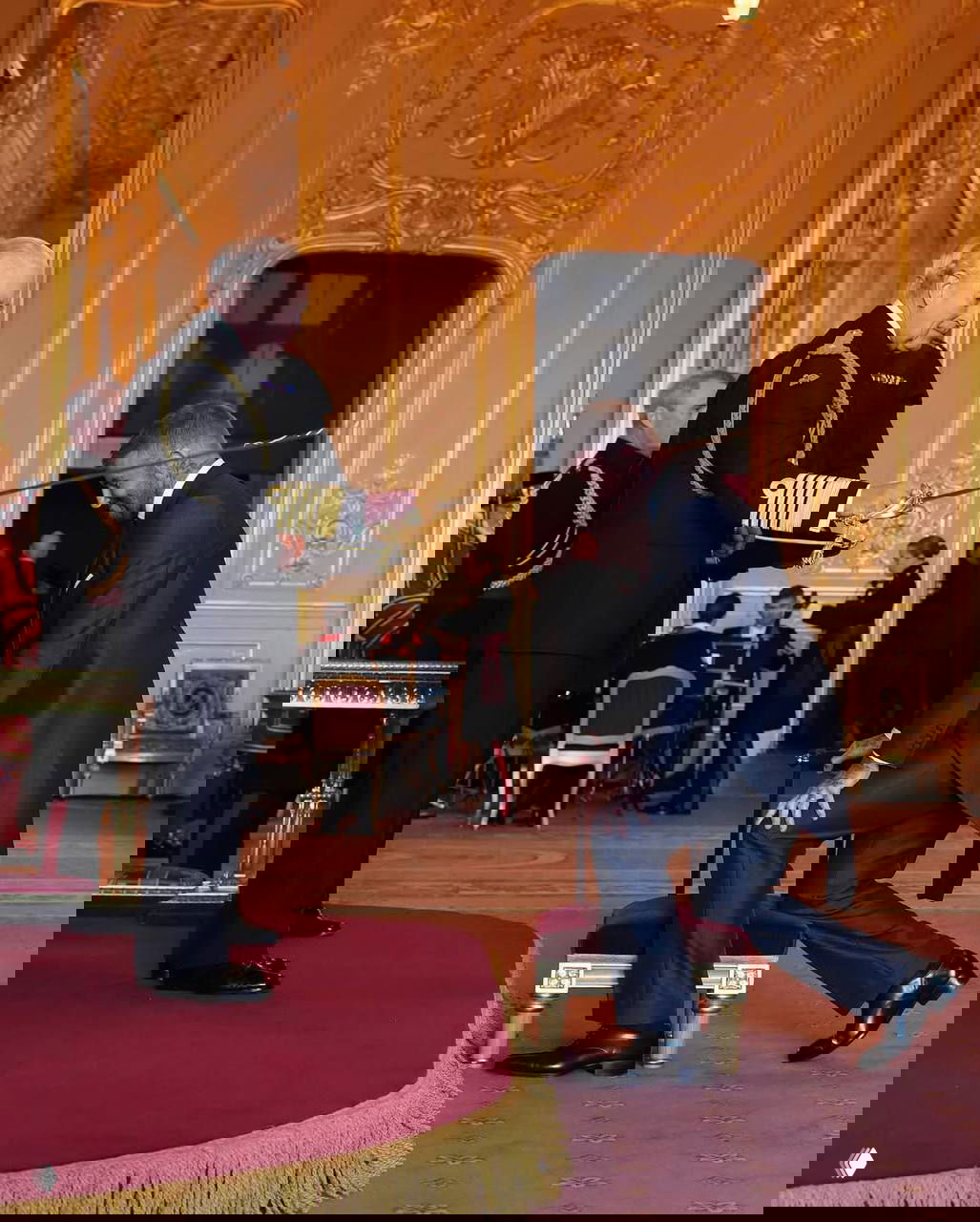 Момент посвящения: король Карл III касается церемониальным мечом плеча Дэвида Бекхэма в официальной обстановке с музыкантами и чиновниками позади.