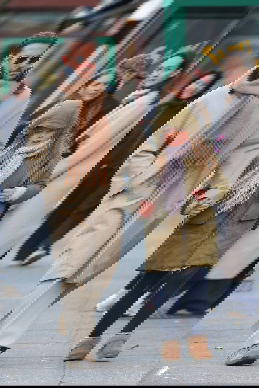 Мужчина в солнцезащитных очках и тёплом шарфе идёт рядом с ребёнком в зелёной куртке, вокруг прохожие на городском тротуаре.