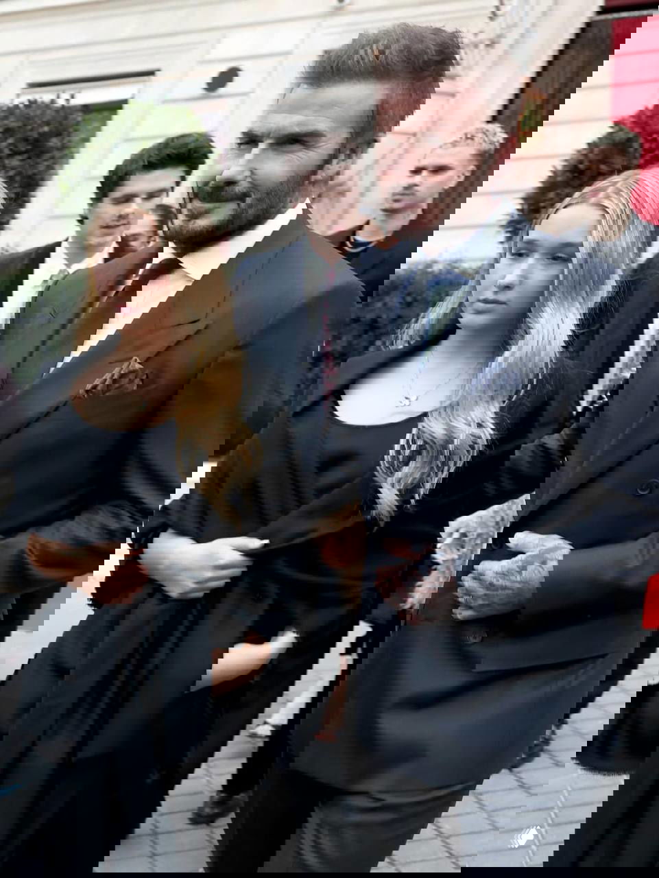 Мужчина в тёмном костюме идёт рядом с молодой девушкой в чёрном платье; кадр сделан у входа на мероприятие в Париже при дневном свете.