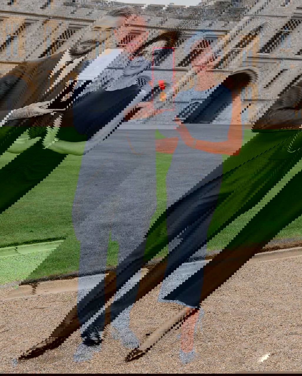 Дэвид и Виктория Бекхэм на лужайке у Виндзорского замка: он держит коробочку с медалью, она в элегантном платье и вуалетке, пара позирует фотографам.