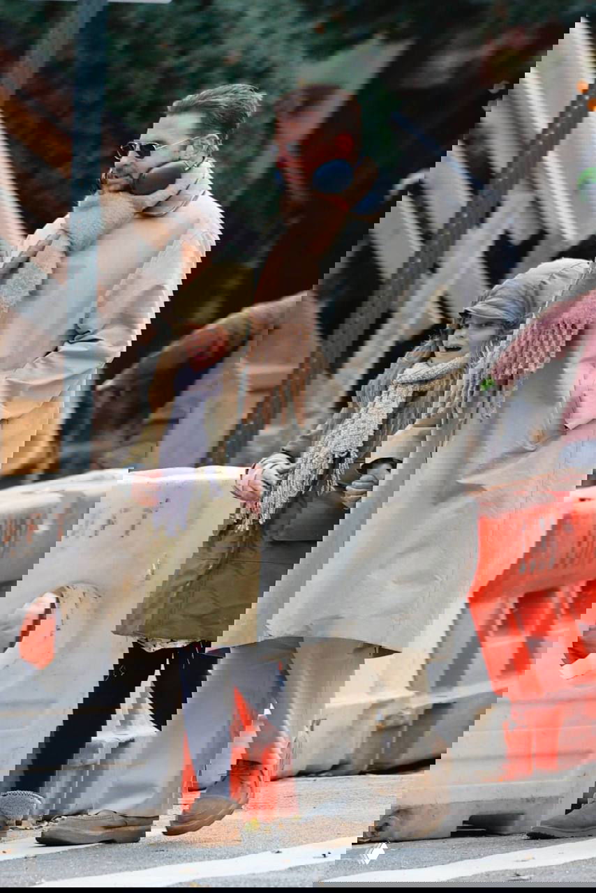 Мужчина в бежевом плаще и ребёнок в зелёной парке на фоне городских барьеров и уличной сцены во время прогулки.