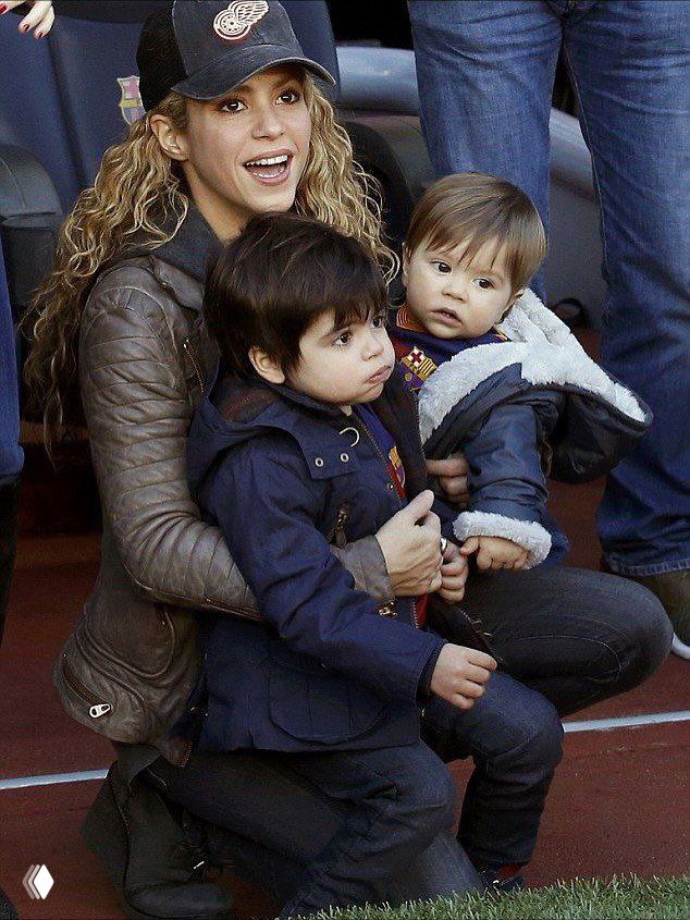 Женщина на спортивной трибуне держит младшего ребёнка на коленях, рядом старший сын, атмосфера матча и семейная поддержка.