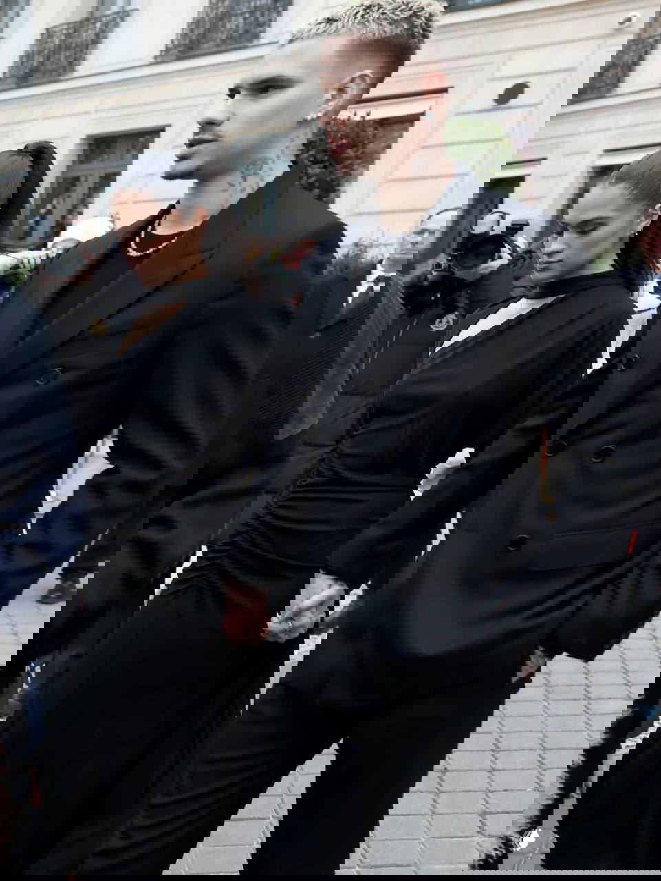 Молодой мужчина с короткой стрижкой и девушка в чёрном платье держатся за руки и идут по тротуару на фоне фотографов на мероприятии в Париже.