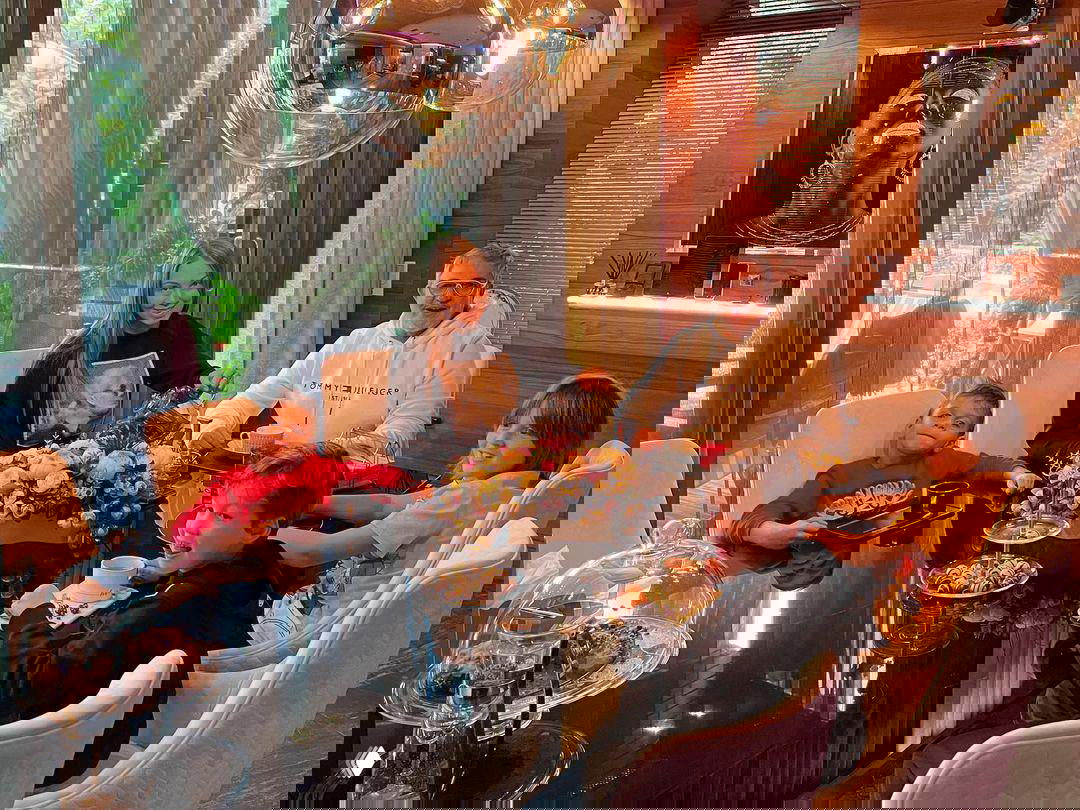 Мужчина и четверо детей за столом в элегантной столовой: сервировка с чашками, сладостями и цветочной композицией на стеклянной поверхности стола.