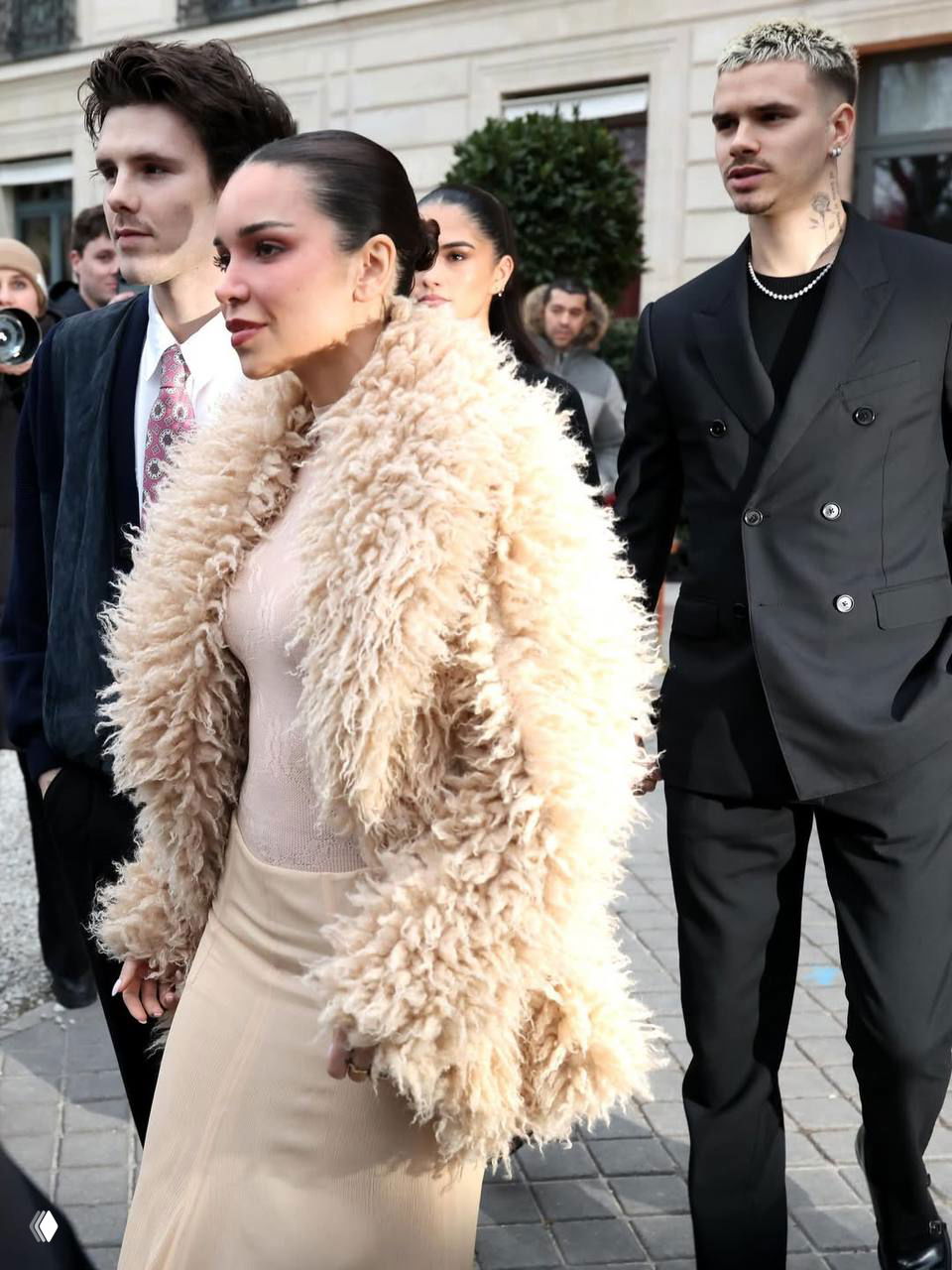 Группа гостей модного показа: женщина в пушистой бежевой шубе и молодые люди в элегантных чёрных образах идут по улице перед входом в зал.