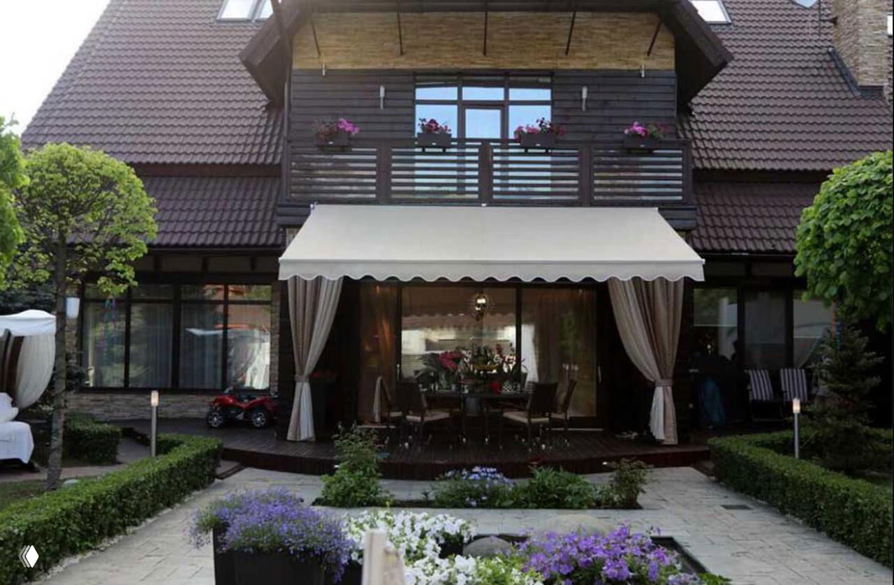 Фасад загородной виллы со стороны сада: двухэтажный дом с террасой, маркизой, украшенными цветами балконами и аккуратно ухоженной дорожкой.