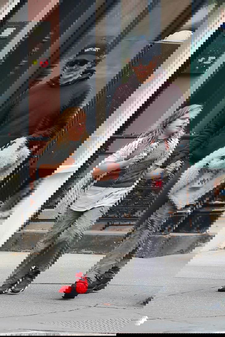 Папарацци‑кадр: мужчина в тёмной толстовке держит за руку девочку с мороженым на городском тротуаре, уличная сцена и витрины зданий вокруг.