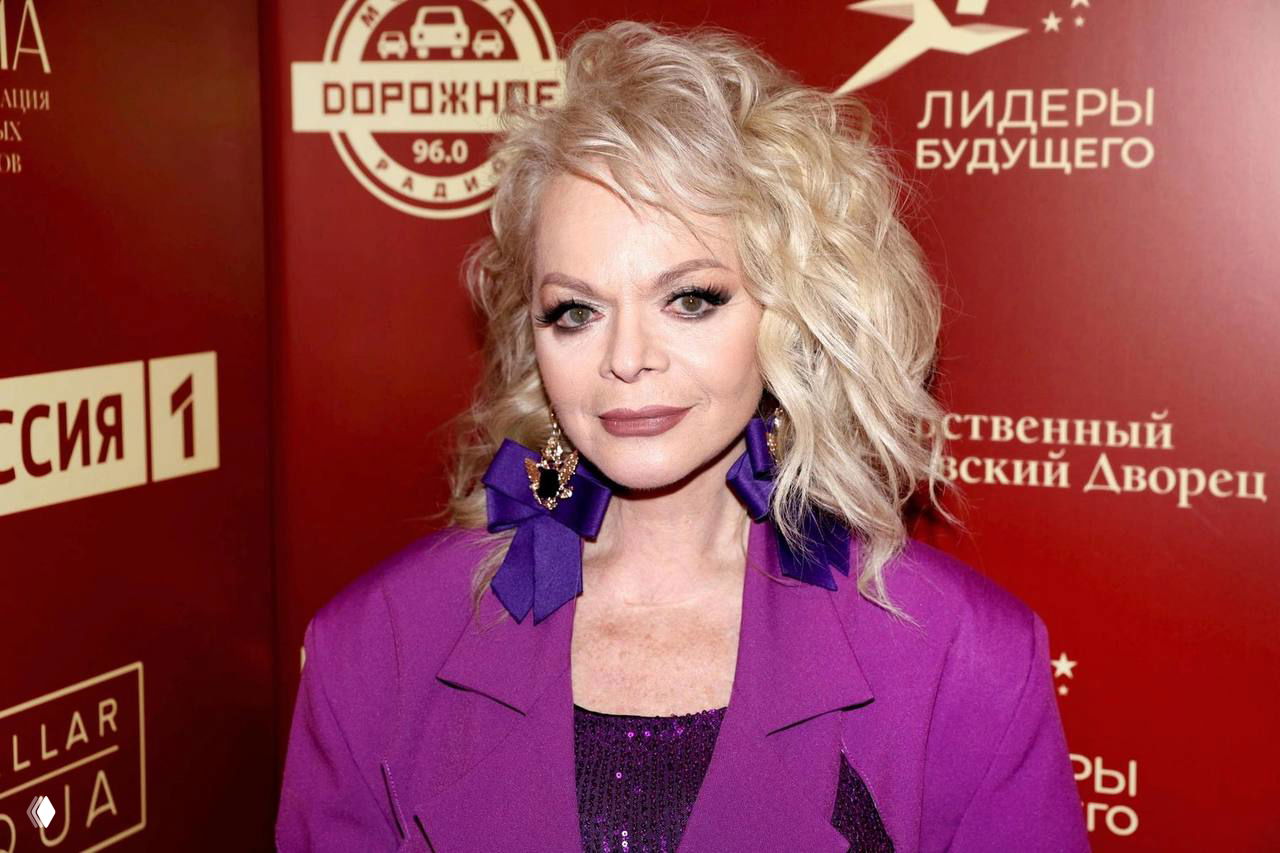 Женщина в фиолетовом пиджаке и крупными серьгами-бантиками на красной фотозоне с логотипами, портретный план, пресс‑мероприятие.