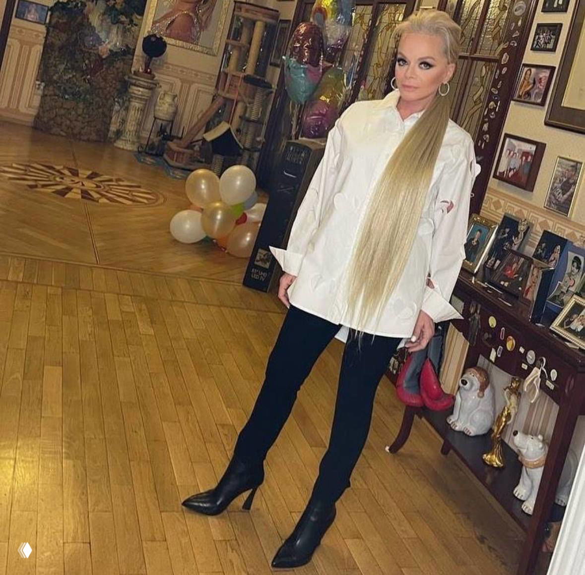 Женщина в белой рубашке и высоких сапогах позирует в парадном зале с воздушными шарами и стеной с фоторамками на заднем плане.