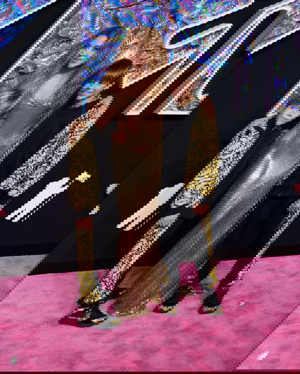 Женщина в золотом платье позирует на красной дорожке вместе с двумя мальчиками в чёрно-золотых нарядах, официальный светский снимок на премьере.