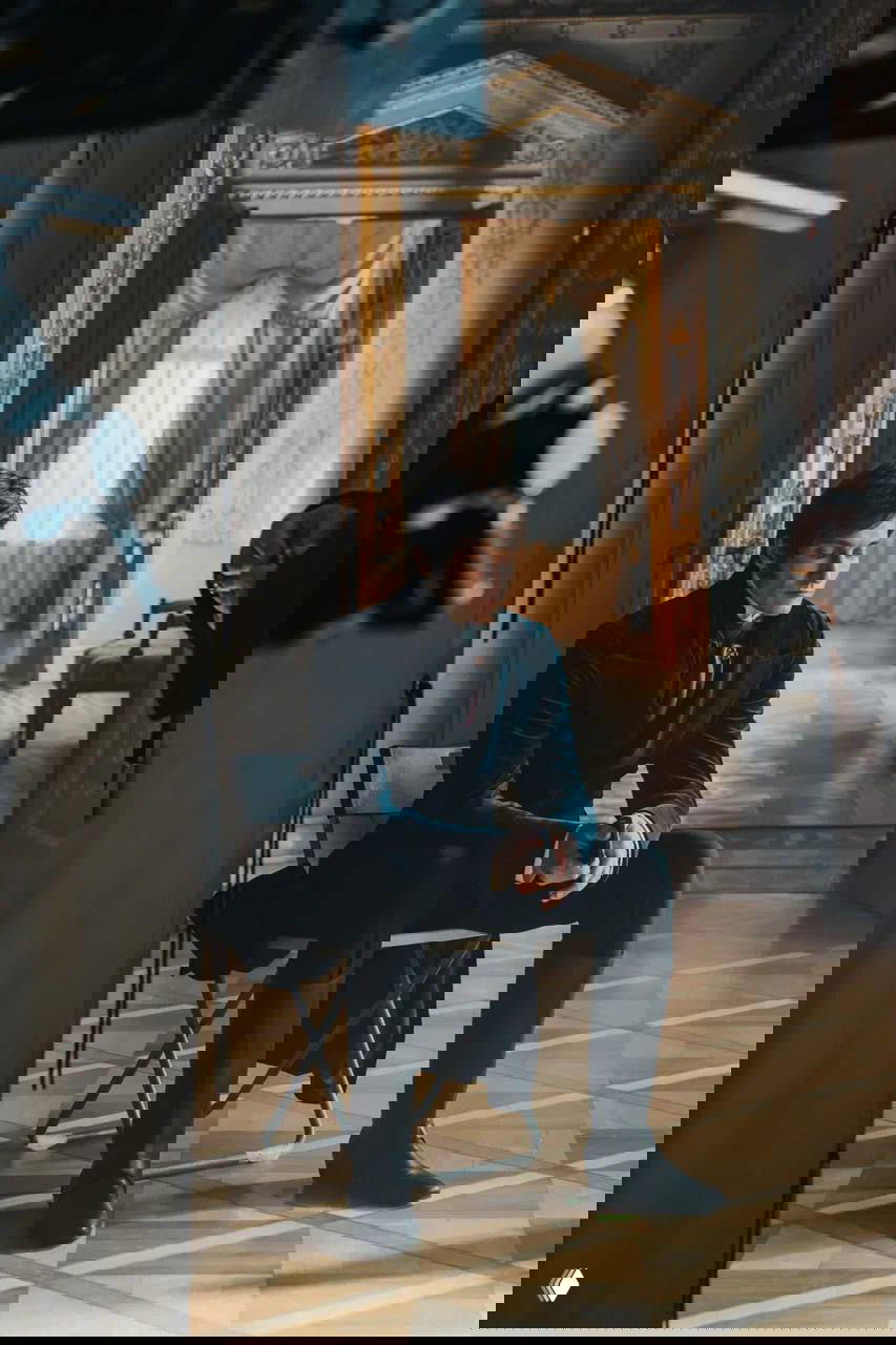 Матвей Лыков сидит на складном кресле между декорациями дворца, задумчиво опирается на руки в сценическом костюме; в кадре видны детали пола и окон.