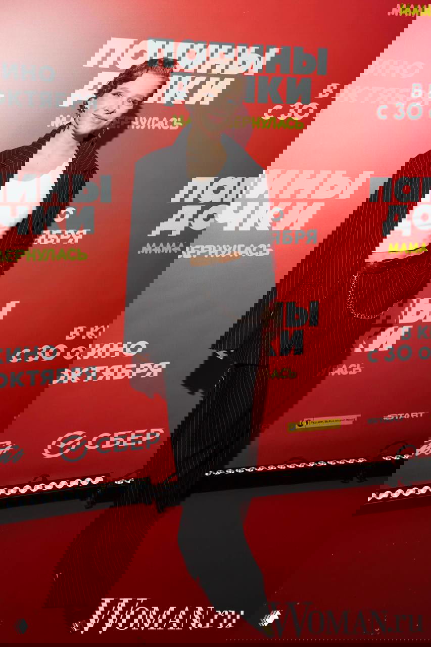 Женщина в свободном тёмном пиджаке и широких брюках позирует на красной дорожке у постера фильма «Папины дочки. Мама вернулась», с логотипом Woman.ru на фото.