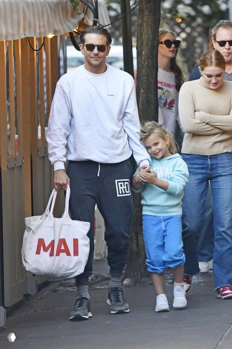 Папарацци‑кадр: мужчина в светлом свитшоте идёт по тротуару, держит за руку маленькую девочку, вокруг прохожие и уличные кафе.