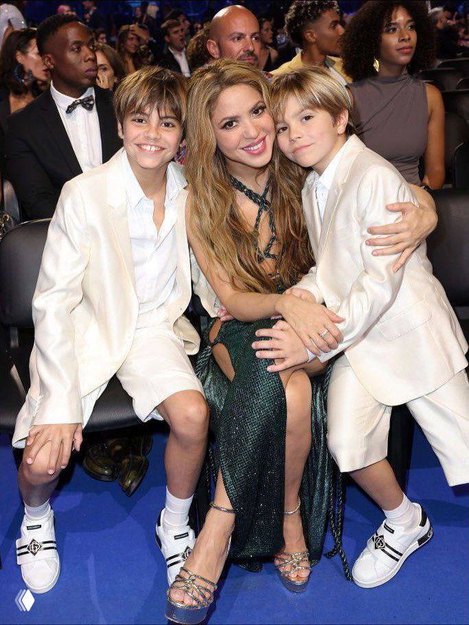 Семейный кадр на церемонии: женщина в тёмном платье сидит между двумя мальчиками в светлых костюмах, радостные эмоции в зале.