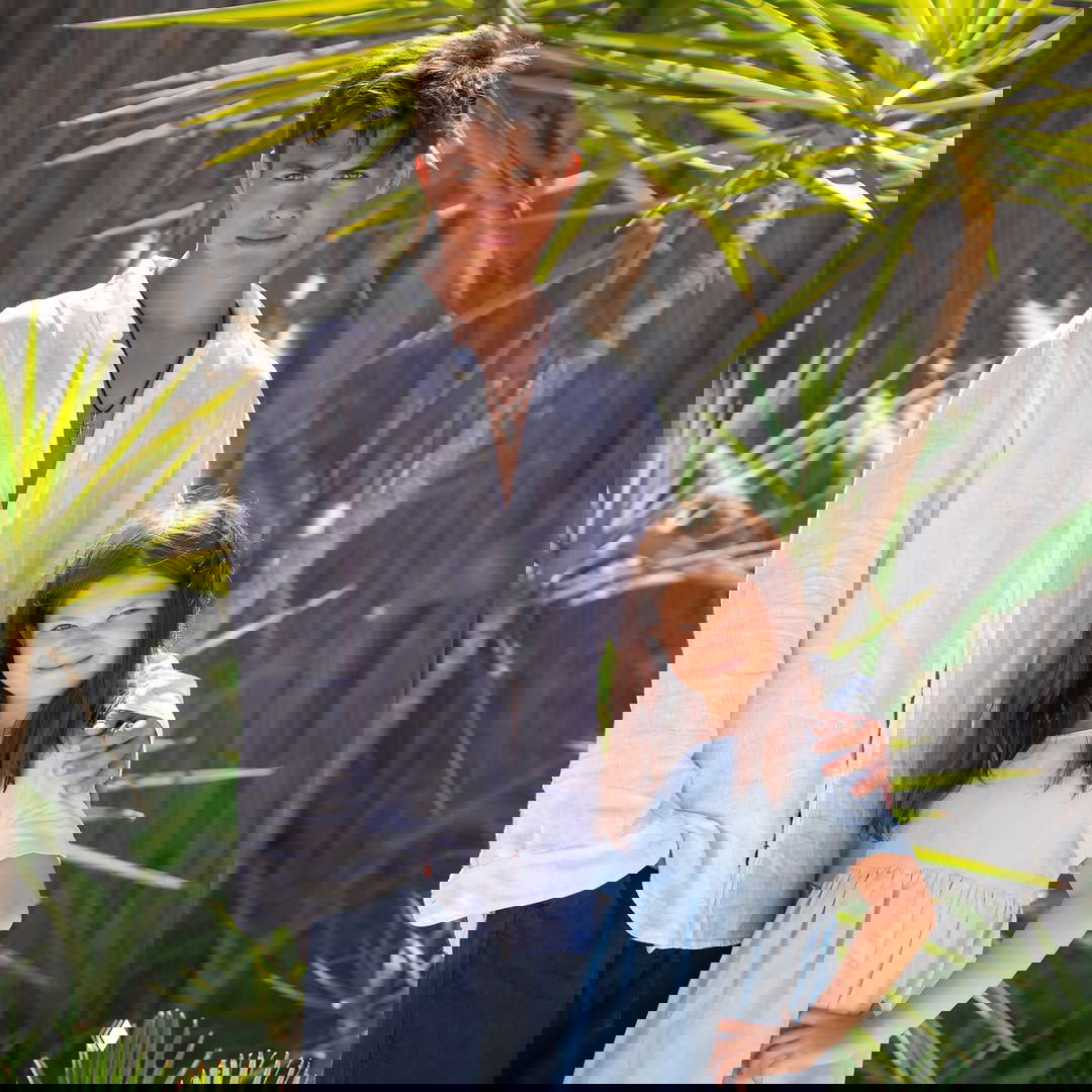 Матвей и младшая доченька стоят на пляжной территории среди пальм и кустарников: тёплые оттенки и семейная сцена на солнце.