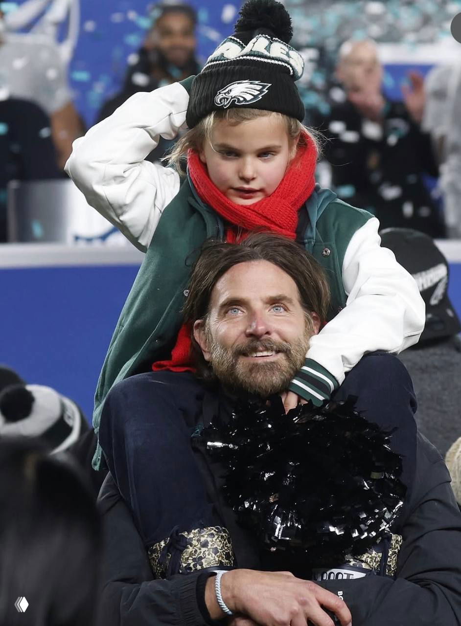 Крупный кадр: мужчина сидит с ребёнком на плечах на массовом мероприятии; на заднем плане городская сцена и праздничная атмосфера.