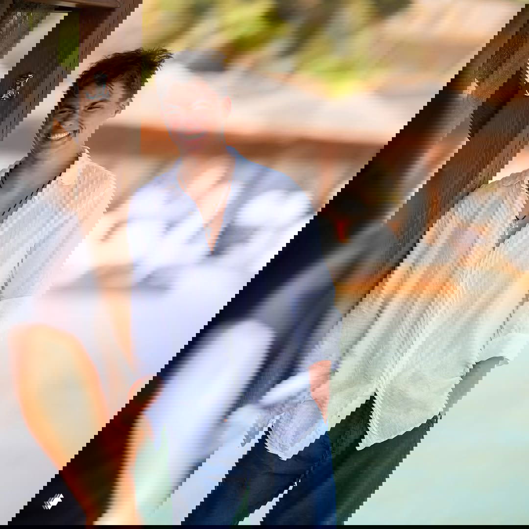 Матвей смеётся в расслабленной позе возле деревянной конструкции на пляже, лёгкая летняя одежда и солнечный свет создают непринуждённый кадр.