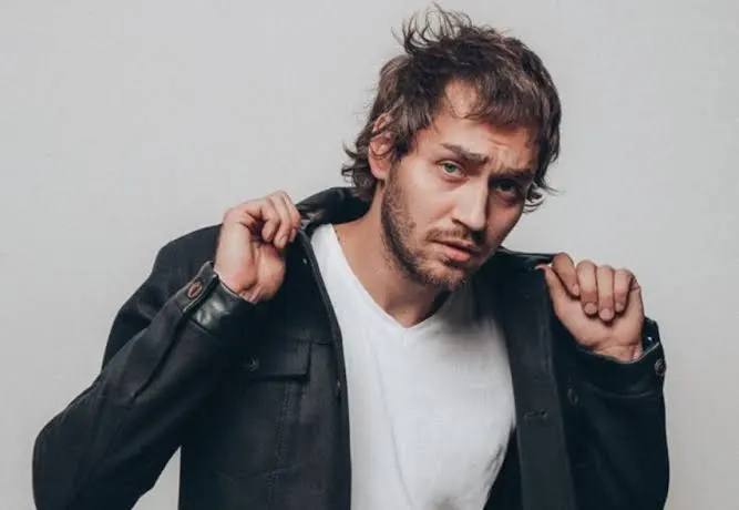 Портрет Рождена Ануси — украинского певца и саунд‑продюсера, в повседневной рубашке и кожаной куртке на нейтральном фоне.