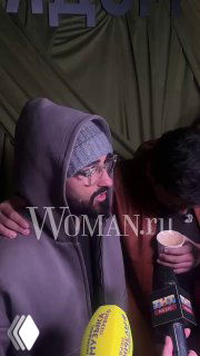 Мужчина в худи и очках стоит на фоне зелёного баннера презентации, посетители держат напитки; кадр из зоны встречи гостей и прессы.