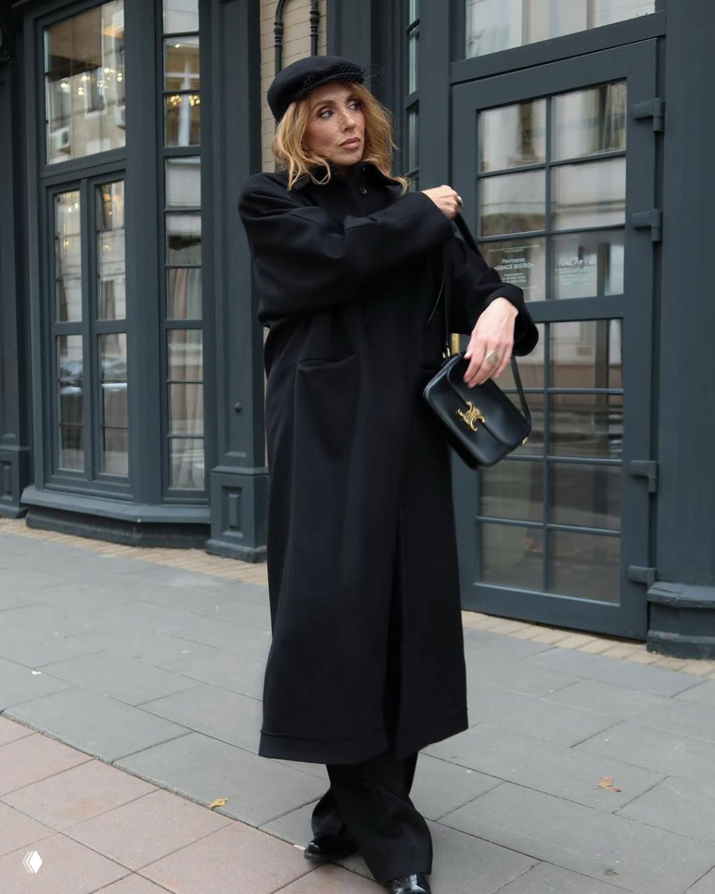 Полный рост: Светлана Бондарчук в длинном черном пальто и сумке, позирует на городской улице у стеклянных витрин, монохромный образ.