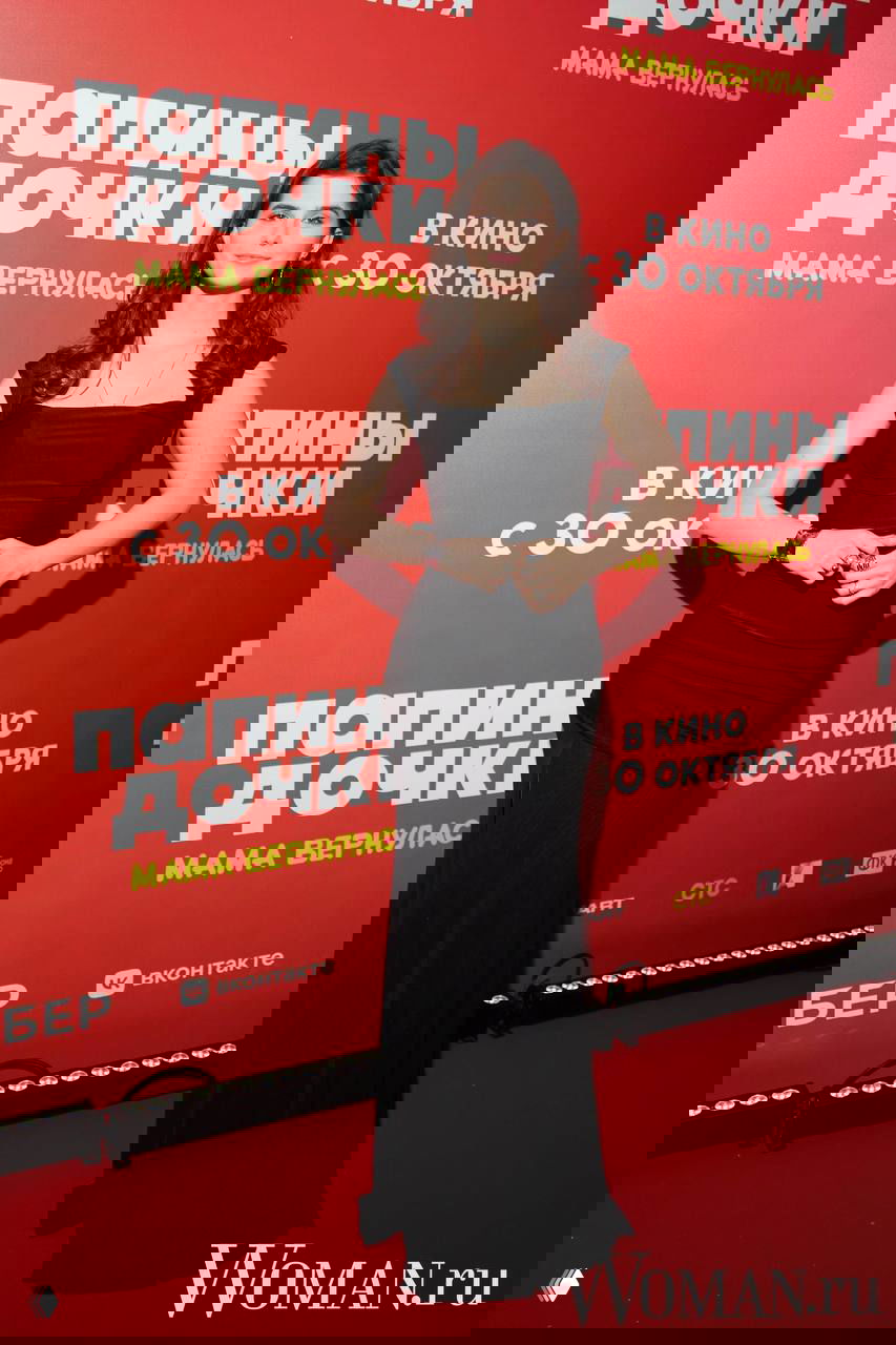 Женщина в элегантном чёрном платье позирует на красной дорожке, на фоне виден логотип и постер фильма «Папины дочки. Мама вернулась», логотип Woman.ru присутствует.