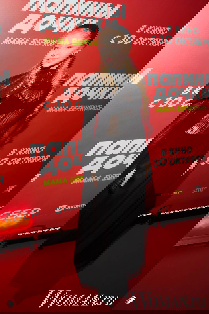 Подросток в чёрном платье и длинной юбке позирует на красной дорожке на фоне постера премьеры «Папины дочки. Мама вернулась», также виден логотип Woman.ru на фото.