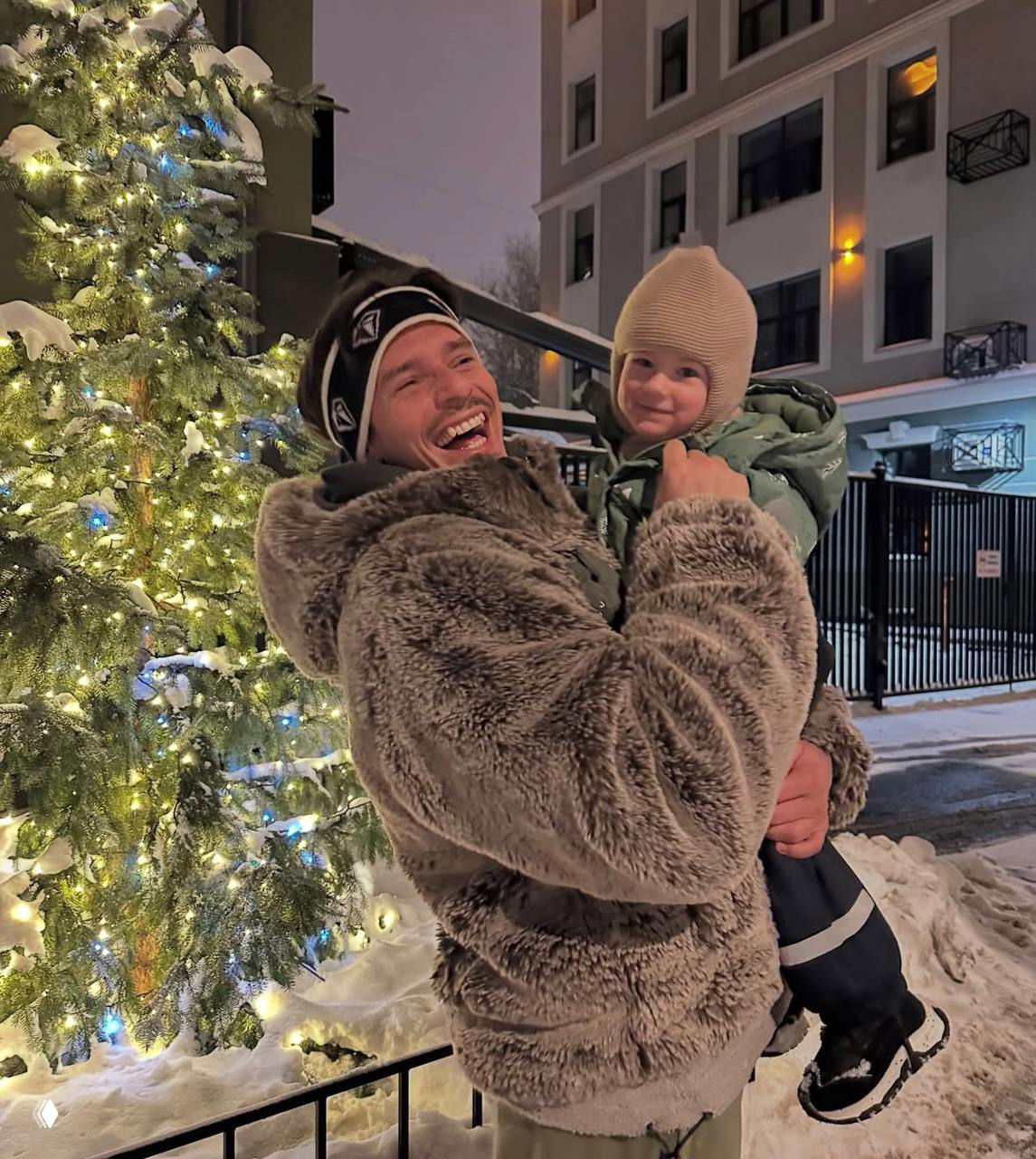 Даниил Наумов с ребёнком у подсвеченной ёлки на фоне жилого дома зимой — семейная сцена, слышны улыбки и радость момента.