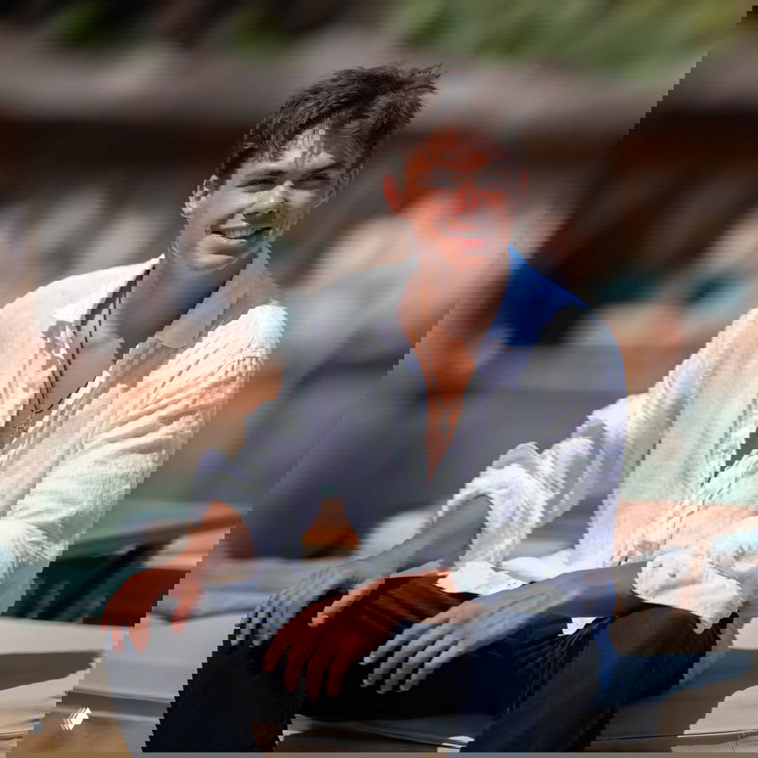 Матвей в белой рубашке сидит у воды в непринуждённой позе, расслабленное выражение лица и мягкий боке на заднем плане.