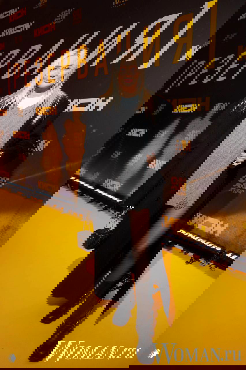 Женщина в чёрном пальто и ботинках на жёлтой ковровой дорожке премьеры; повседневный тёплый образ среди гостей вечера.