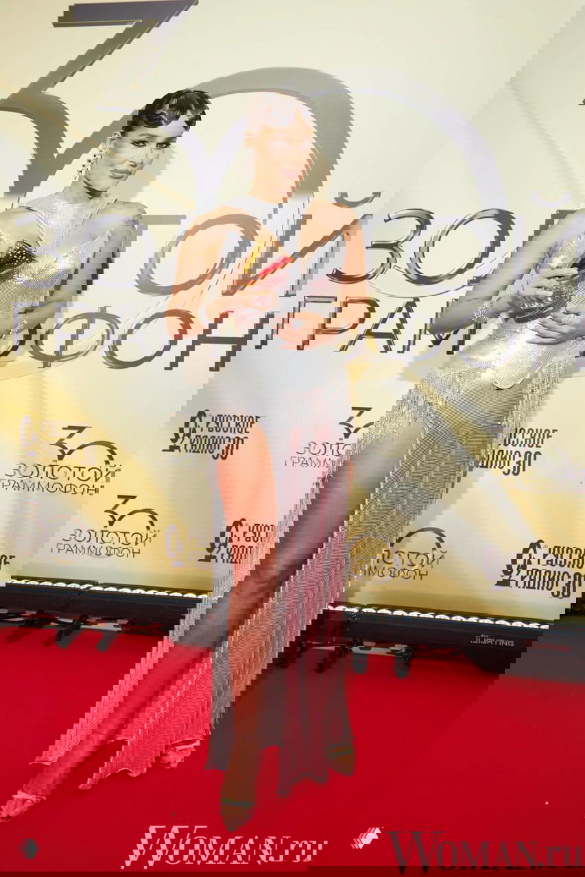 Женщина в серебристом вечернем платье с высоким разрезом, украшениями и статуэткой позирует на красной дорожке у баннера юбилея премии.