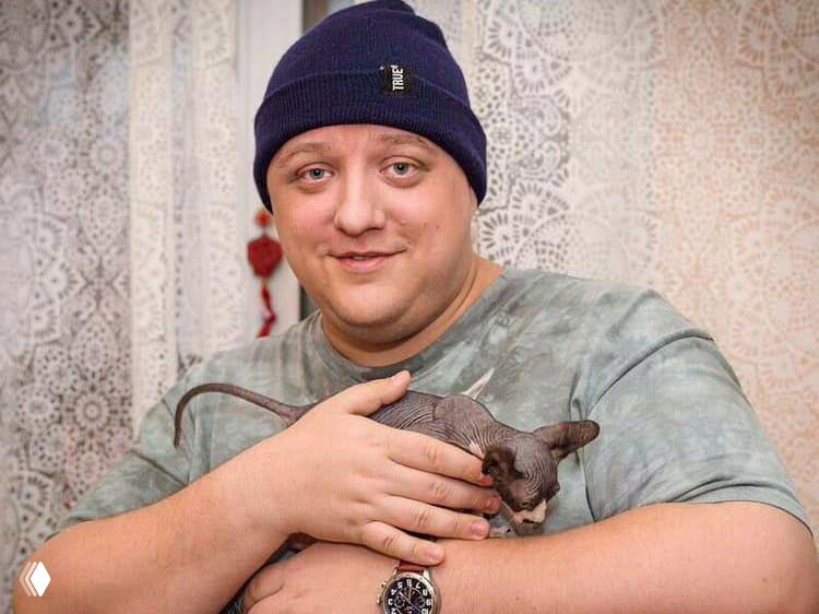 Улыбающийся мужчина в тёмной шапке и футболке держит голого бесшёрстного кота (порода сфинкс) на фоне домашнего интерьера