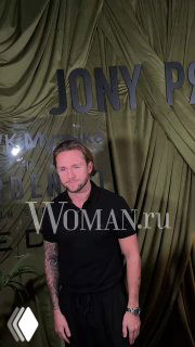 Мужчина в чёрной футболке позирует у зелёного баннера презентации с крупными буквами; кадр из пресс-зоны, водяной знак Woman.ru.