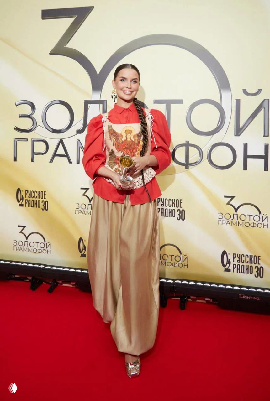 Женщина в красной блузе и широких золотистых брюках держит статуэтку и улыбается на красной дорожке в зоне с логотипами юбилейного «Золотого Граммофона».