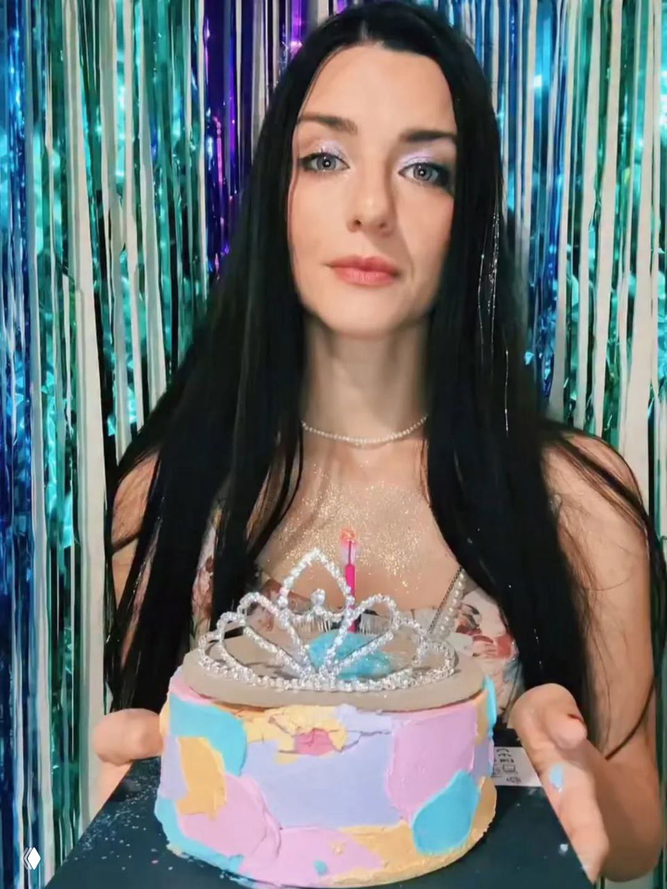 Женщина держит праздничный торт с короной и горящей свечой на фоне блестящих гирлянд — портретный кадр на празднике.