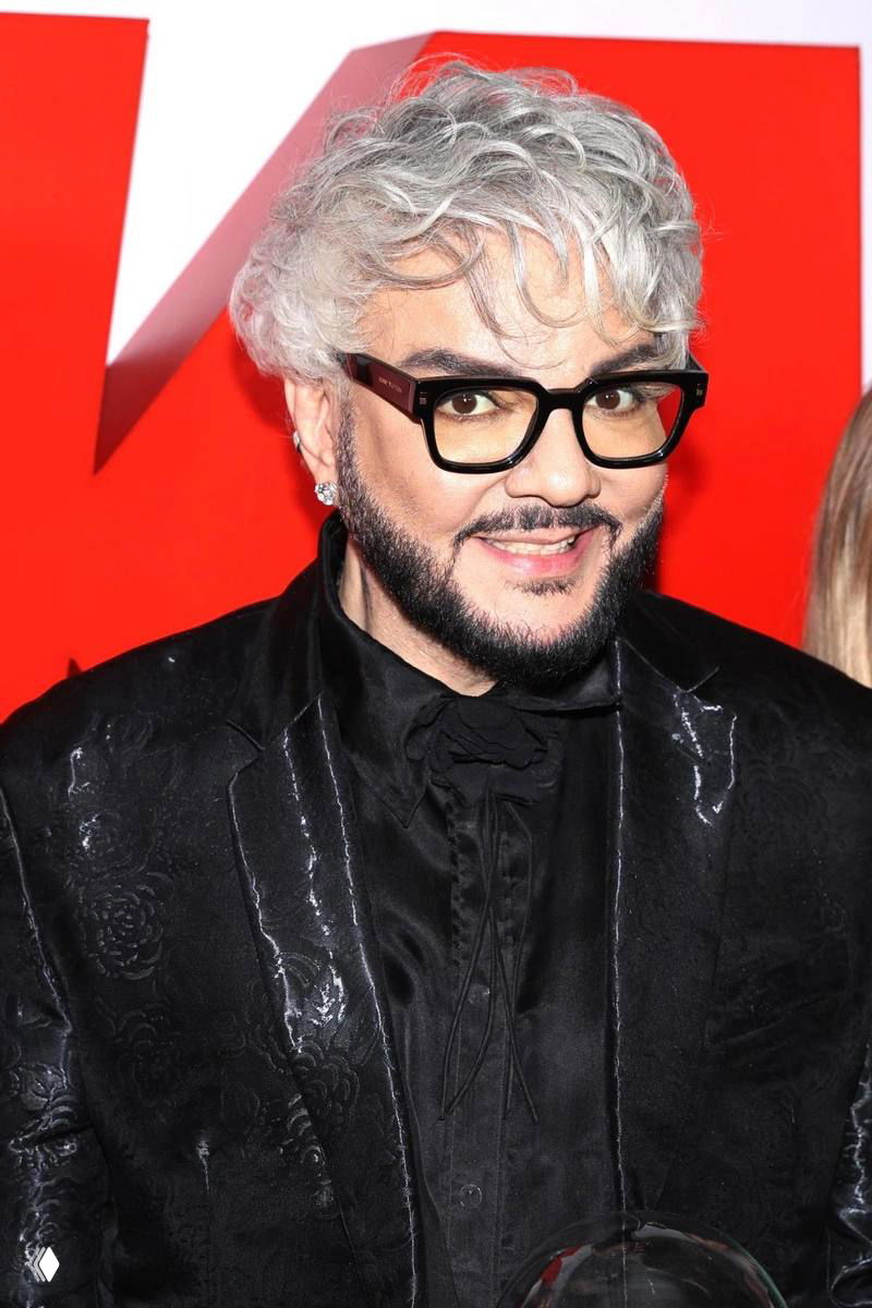 Портретный снимок улыбающегося мужчины в чёрном пиджаке, со светлыми кудрявыми волосами и тёмными очками на красном фоне.