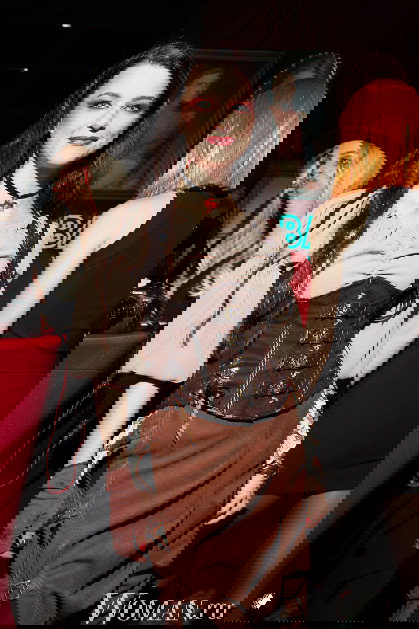 Полноформатный портрет женщины в корсете и юбке на светском мероприятии, вокруг люди и фотокамера на заднем плане.