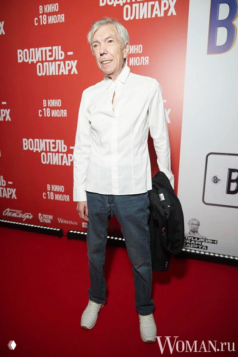 Пожилой мужчина в белой рубашке и джинсах стоит на красной дорожке перед плакатом фильма; на фоне виден логотип Woman.ru.